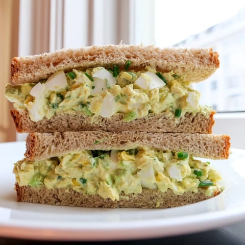 A vibrant sandwich spread featuring mashed avocado egg salad with chives, served on hearty bread with a side of lemon wedges.